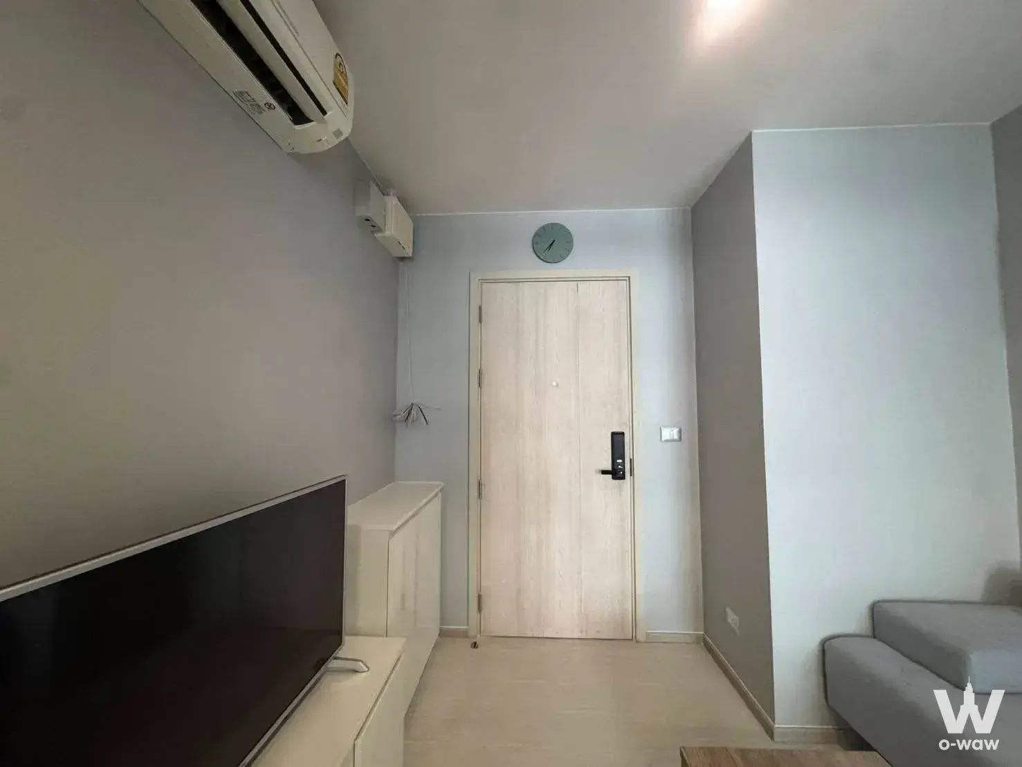 Minimalist living room with beige tiles, white media console, TV, grey sofa, and wood-grain door with smart lock