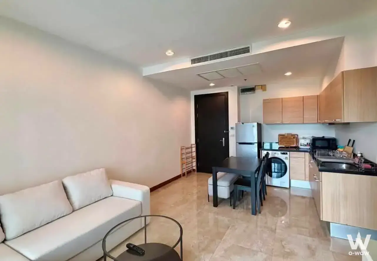 Open-plan living area with beige marble floors, white sofa, and kitchenette in 1-bed condo for rent at The Address Chidlom