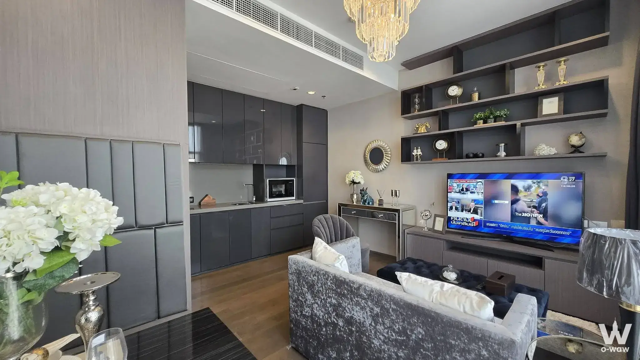 Modern living room with grey velvet sofa, crystal chandelier, and open-plan kitchen in 1-bed rental at The Diplomat Sathorn