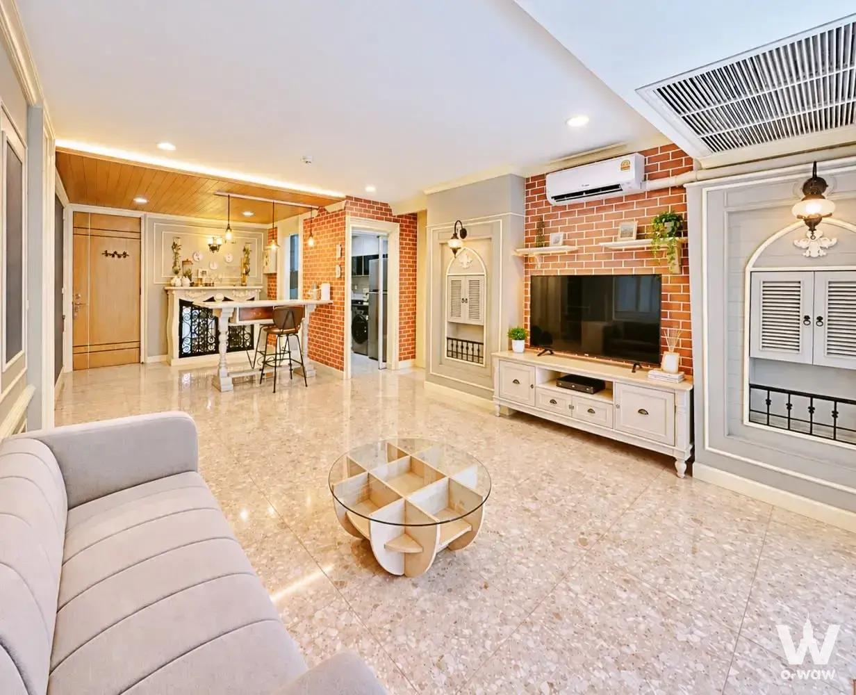 The open-plan living and dining area in a 2-bed Crest Ruamrudee condo has a red brick wall, glass table and terrazzo floor
