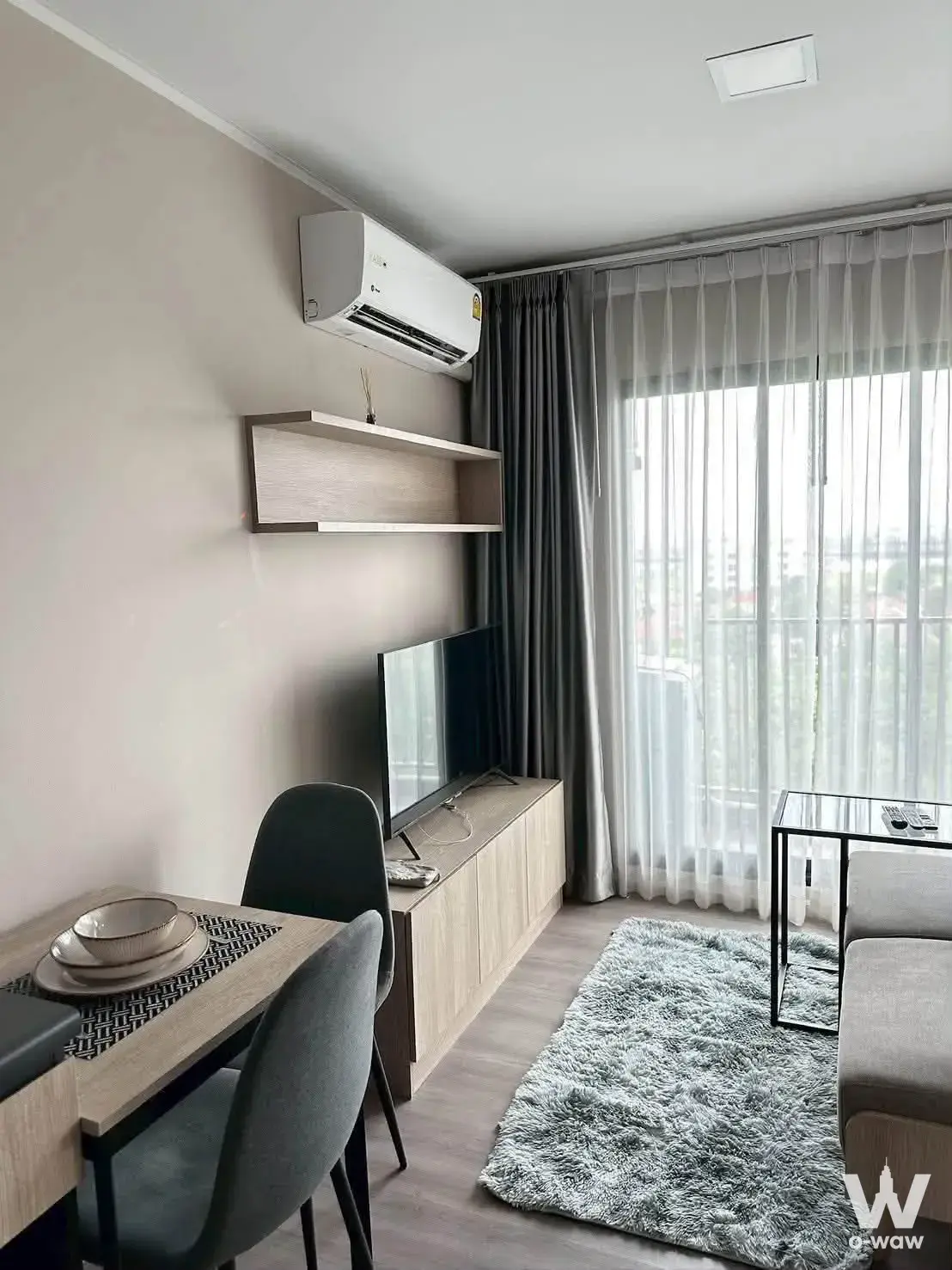 Modern living and dining area with light wood furniture, grey rug, and floor-to-ceiling windows overlooking the city