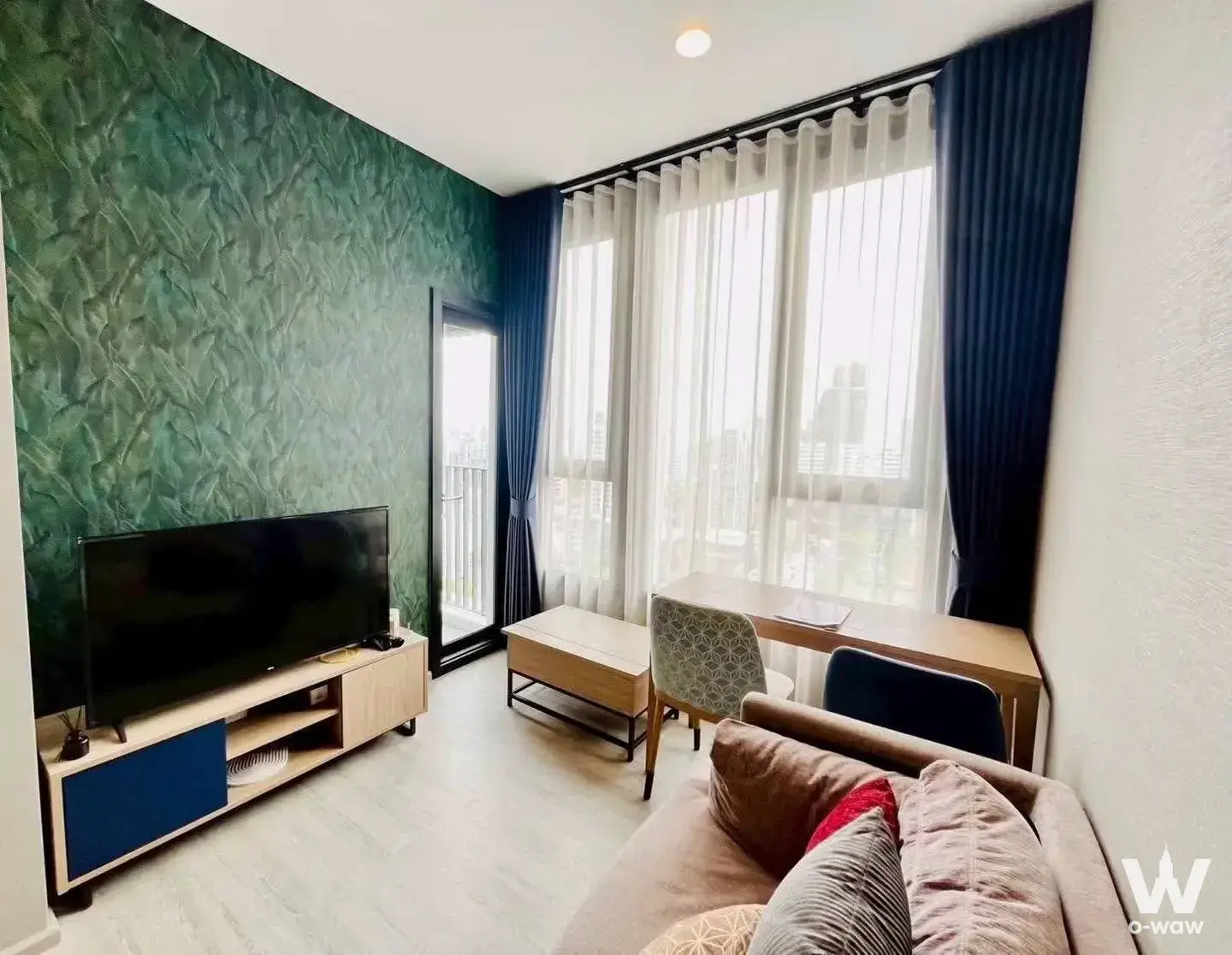 Living room in XT Ekkamai 1-bed condo featuring emerald green accent wall and city view through sheer curtains