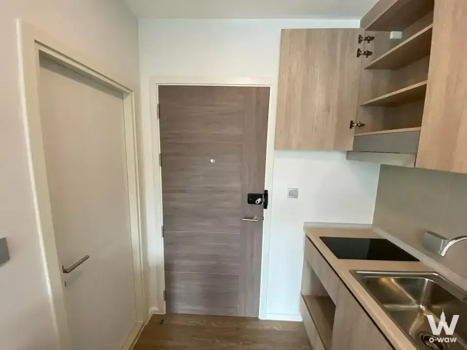 Compact kitchenette with light wood cabinets, induction cooktop, and smart lock entrance in Modiz Sukhumvit 50 condo