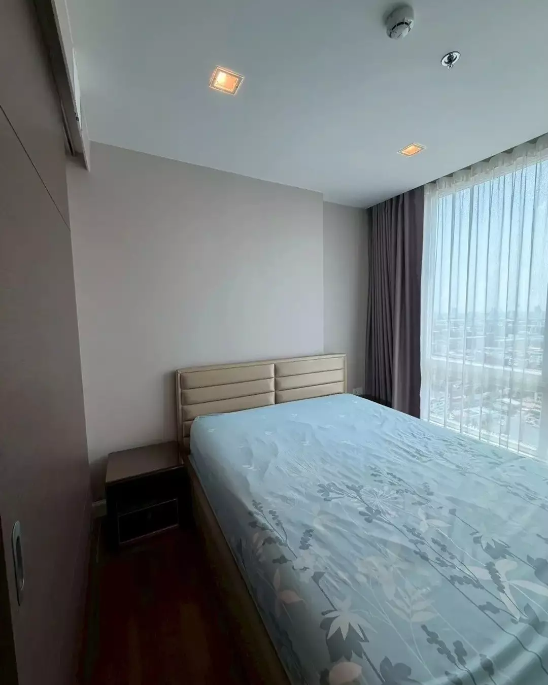 Modern bedroom in 1-bed Polis Condo rental with tufted headboard, blue floral bedding, and large window overlooking Bangkok