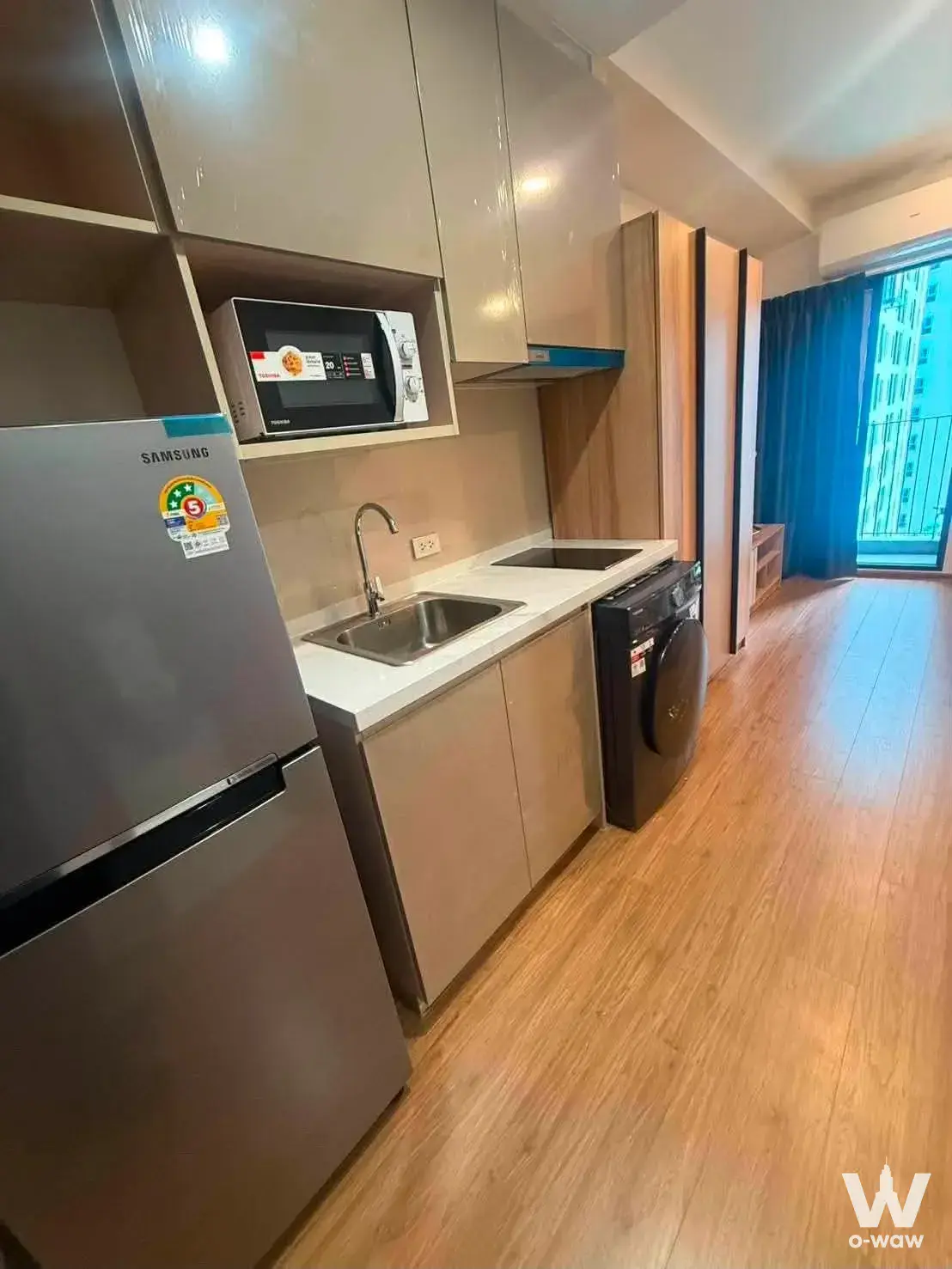 Compact studio kitchen with black induction cooktop, stainless fridge and beige cabinets in The Livin Ramkhamhaeng