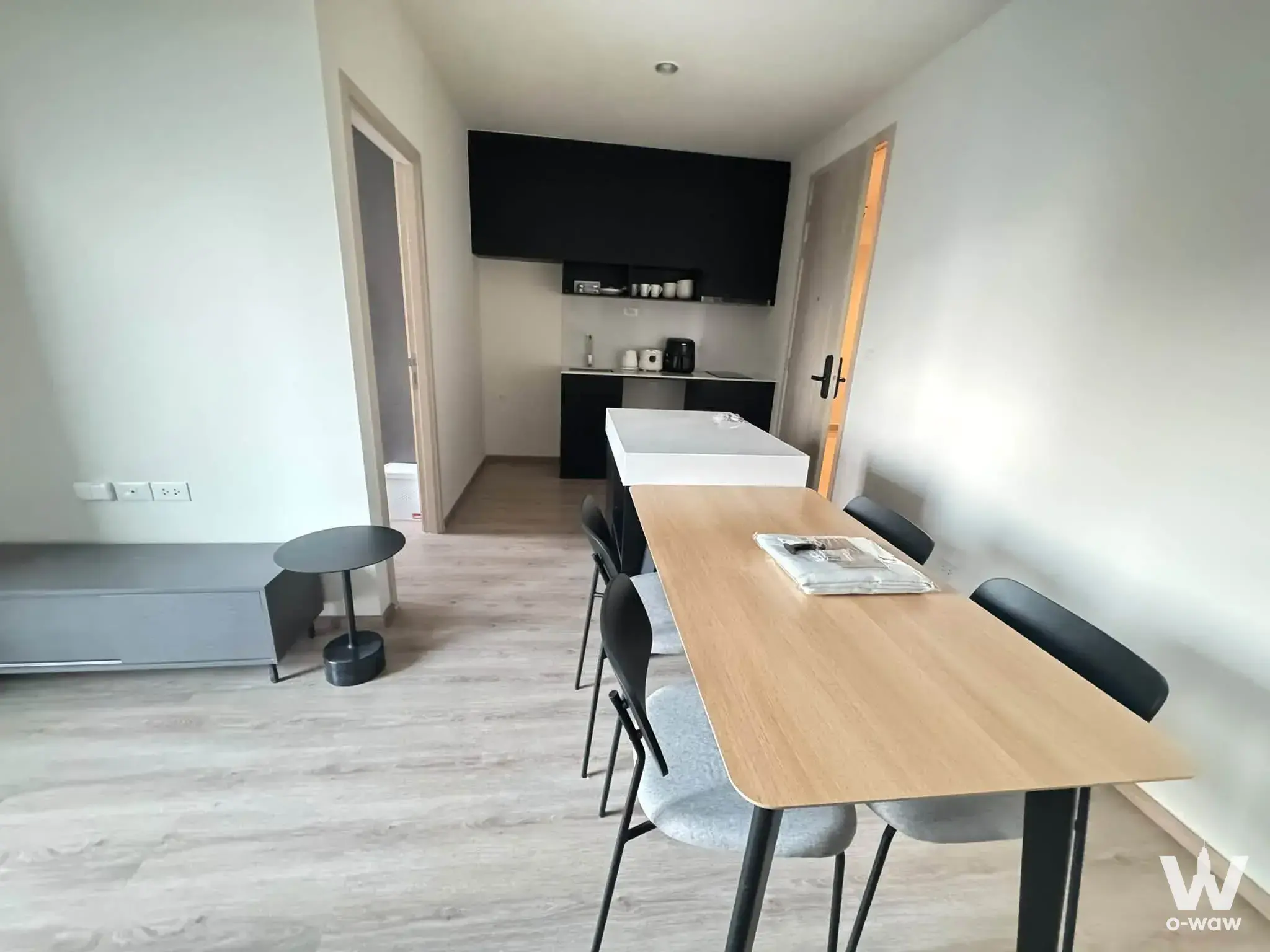This dining area in a 2-bed condo for rent in Nue District R9 has a light wood table, grey chairs and black kitchenette