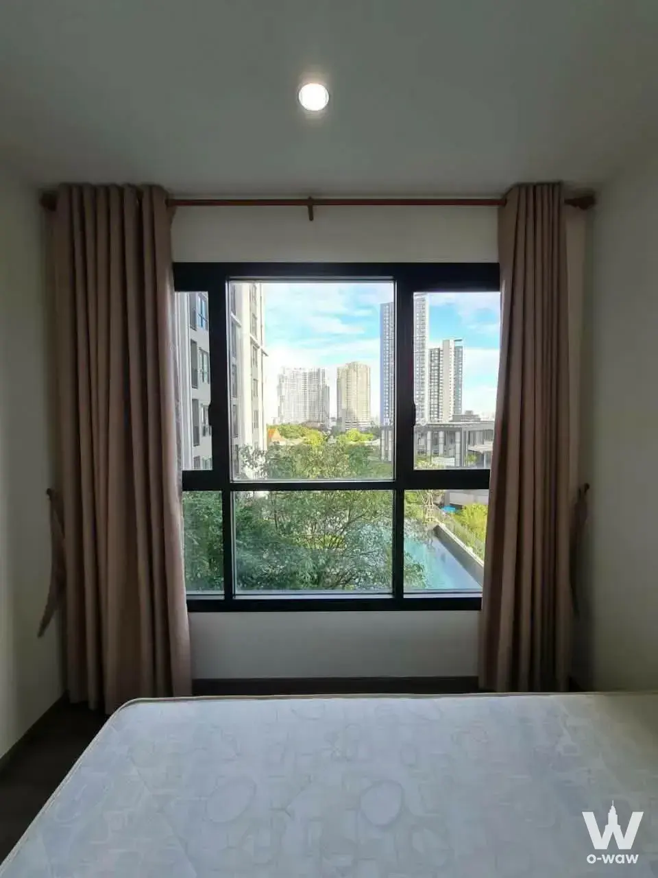 Modern bedroom with white bed, dark wood floors, and large window overlooking trees and high-rises