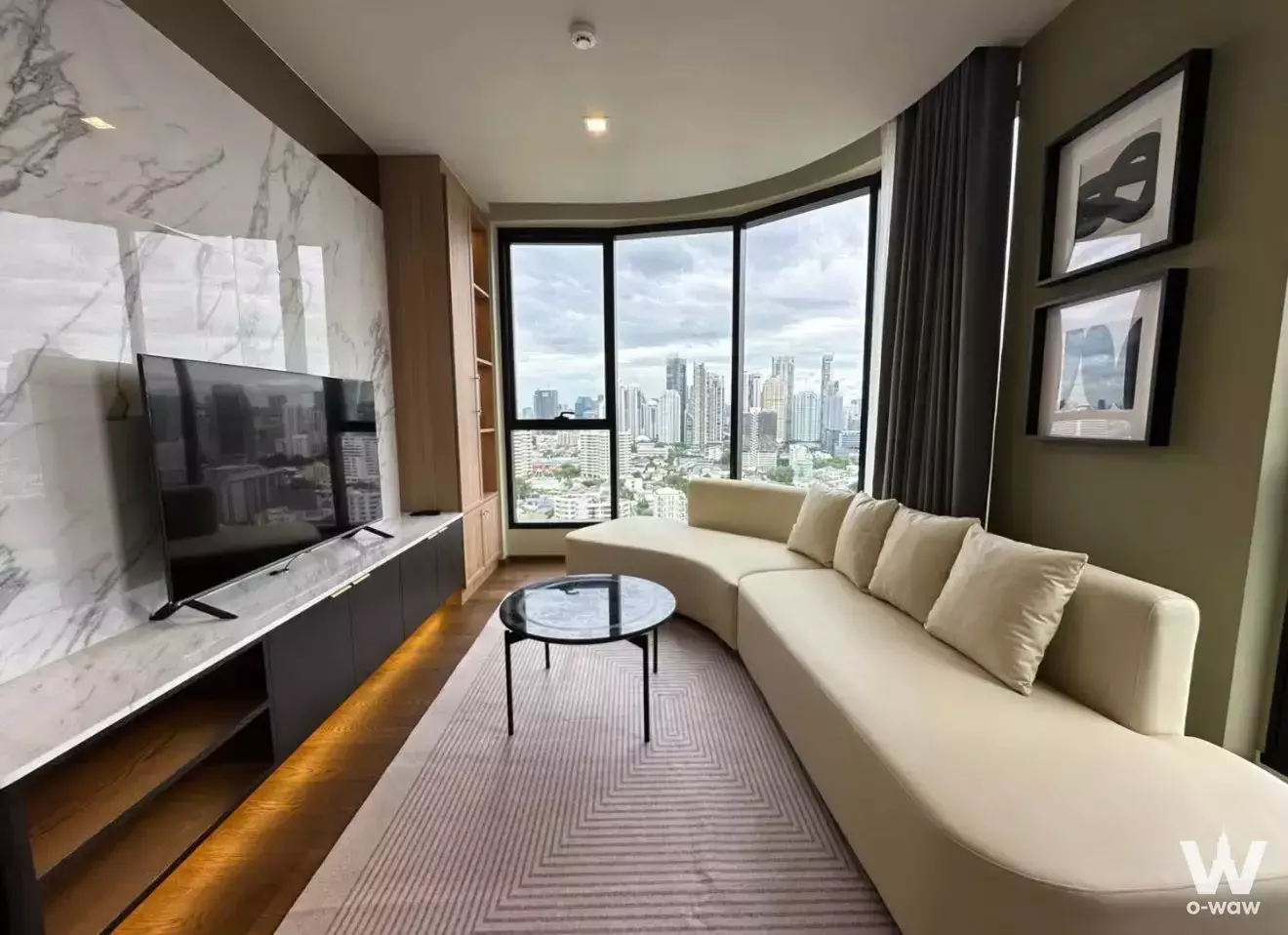 Modern living room with curved windows, city view, cream sofa, and marble TV wall in 2-bed condo for rent at Ideo Q