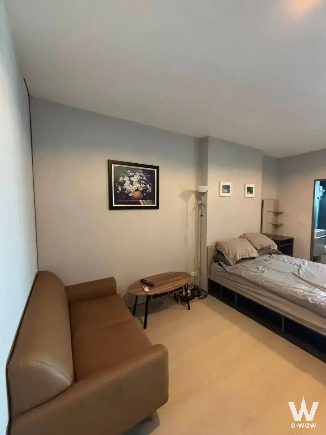 Compact studio with beige sofa, queen bed, and floral artwork in a tidy neutral-toned living space