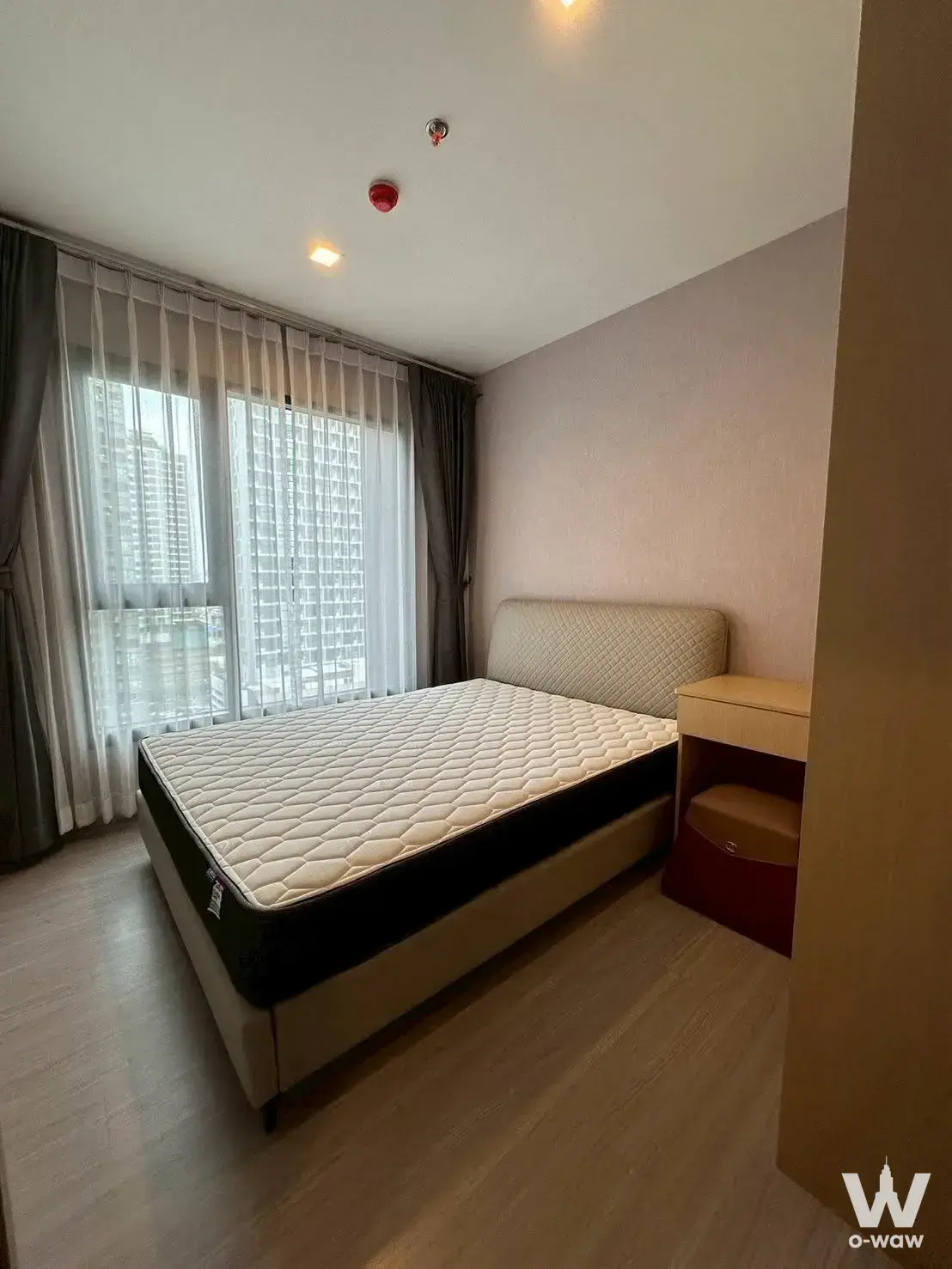 Modern bedroom with light wood flooring, beige bed, and floor-to-ceiling windows offering high-rise city views