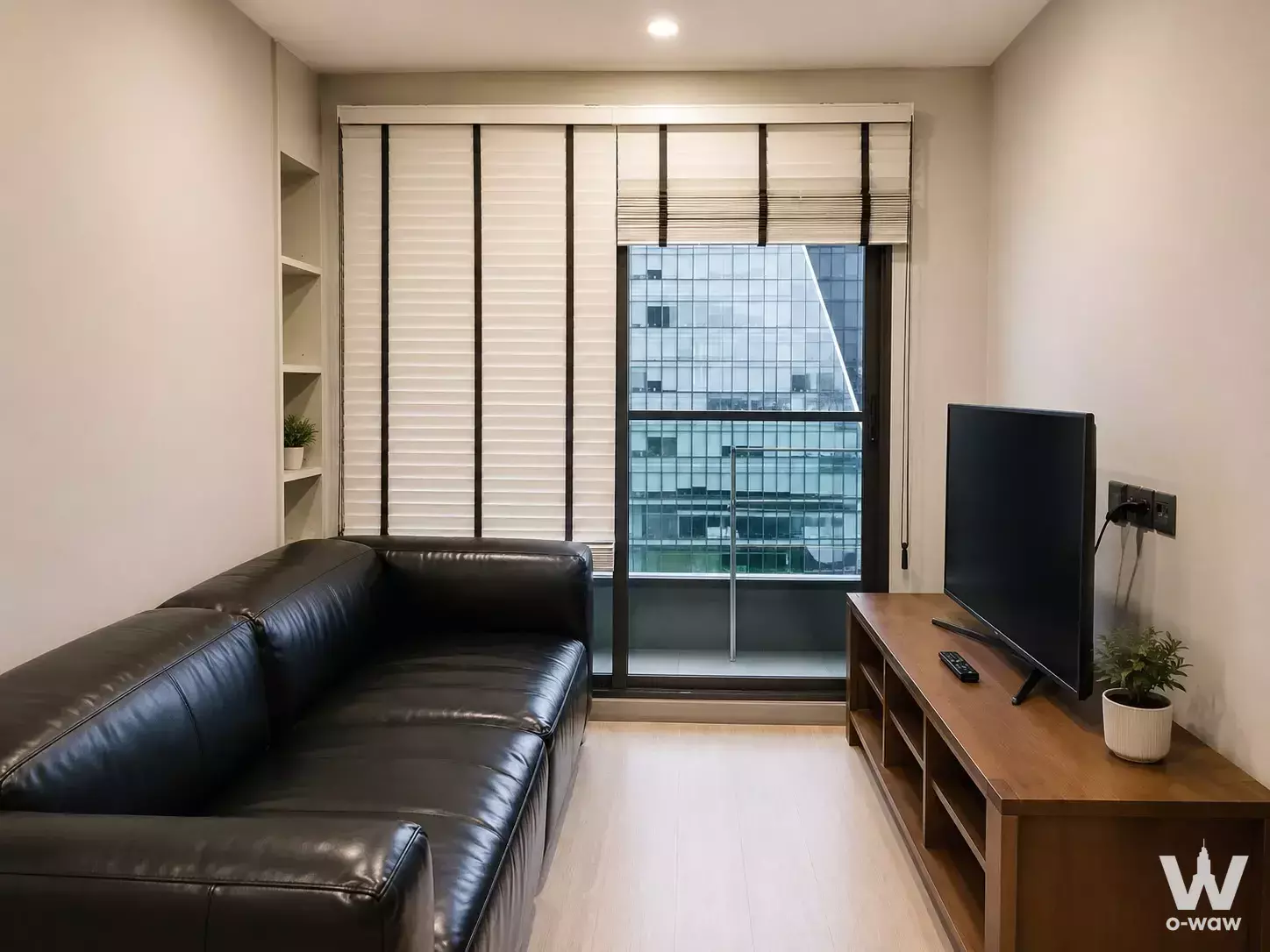 Modern living room in Lumpini Suite condo with black leather sofa, wood media console, and balcony view of city skyline