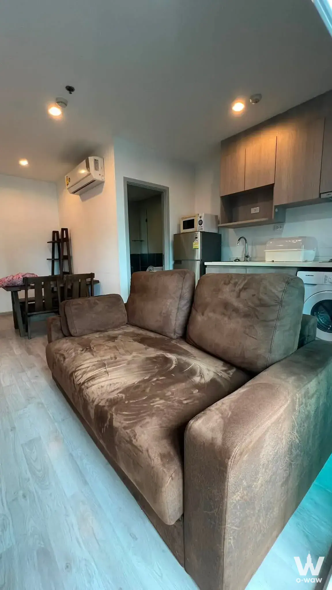 Compact open-plan living space with brown sofa and kitchenette in a 2-bed condo for rent at Ideo Mobi Charan