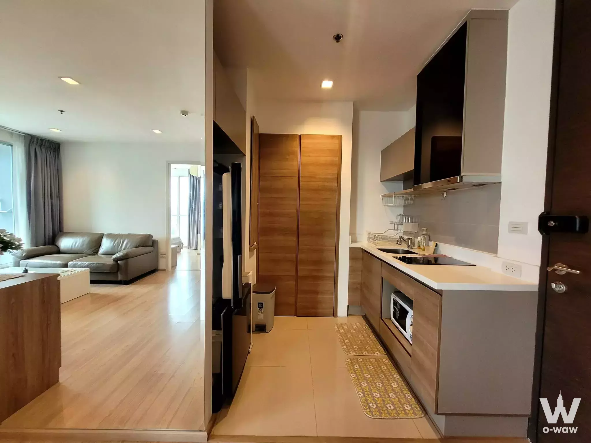Modern open-plan living area with light wood floors, grey sofa, and oak kitchen in 2-bed condo at RHYTHM Phahon-Ari