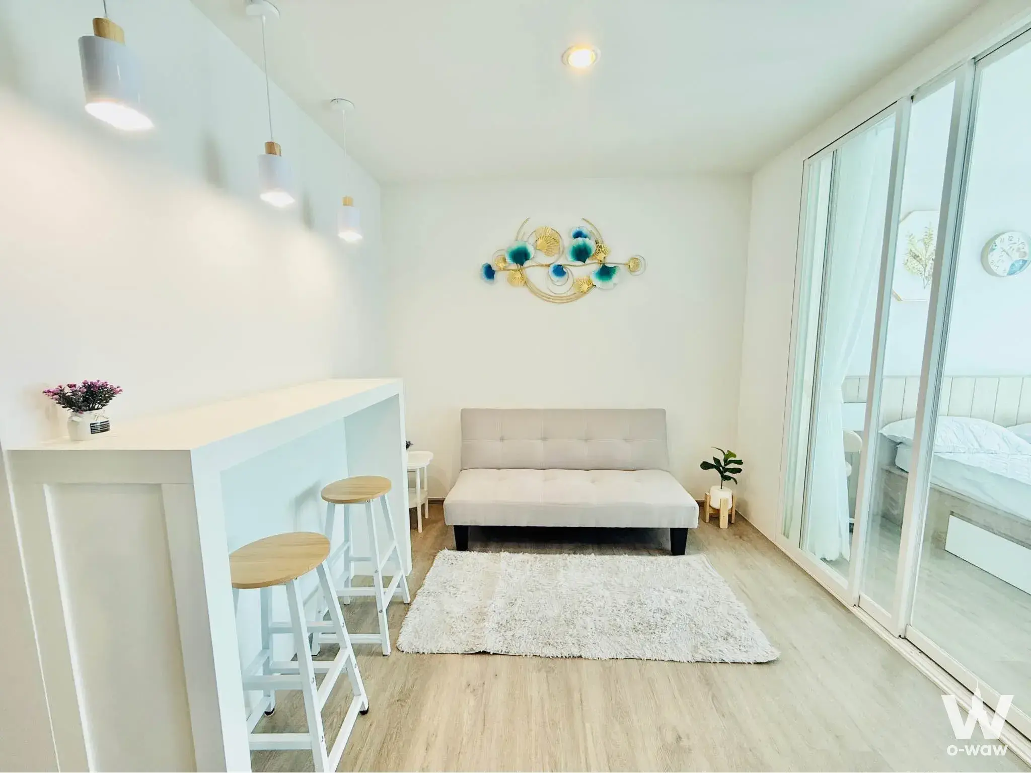 Bright modern studio apartment with light wood floors, beige futon, breakfast bar, and sliding glass doors leading to bedroom