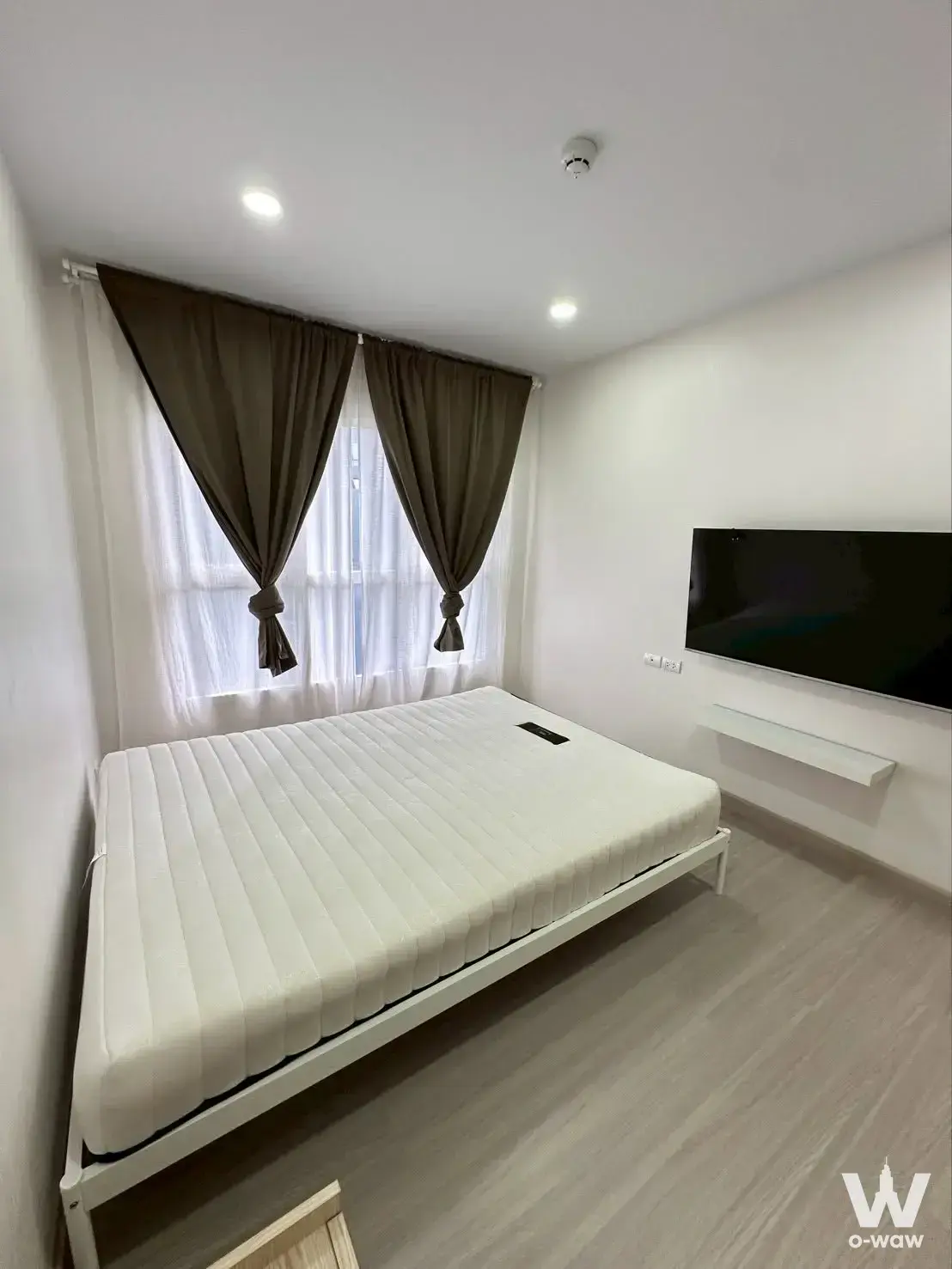 Minimalist bedroom with grey laminate floors, white bed frame, wall-mounted TV, and sheer curtains in Supalai City Resort