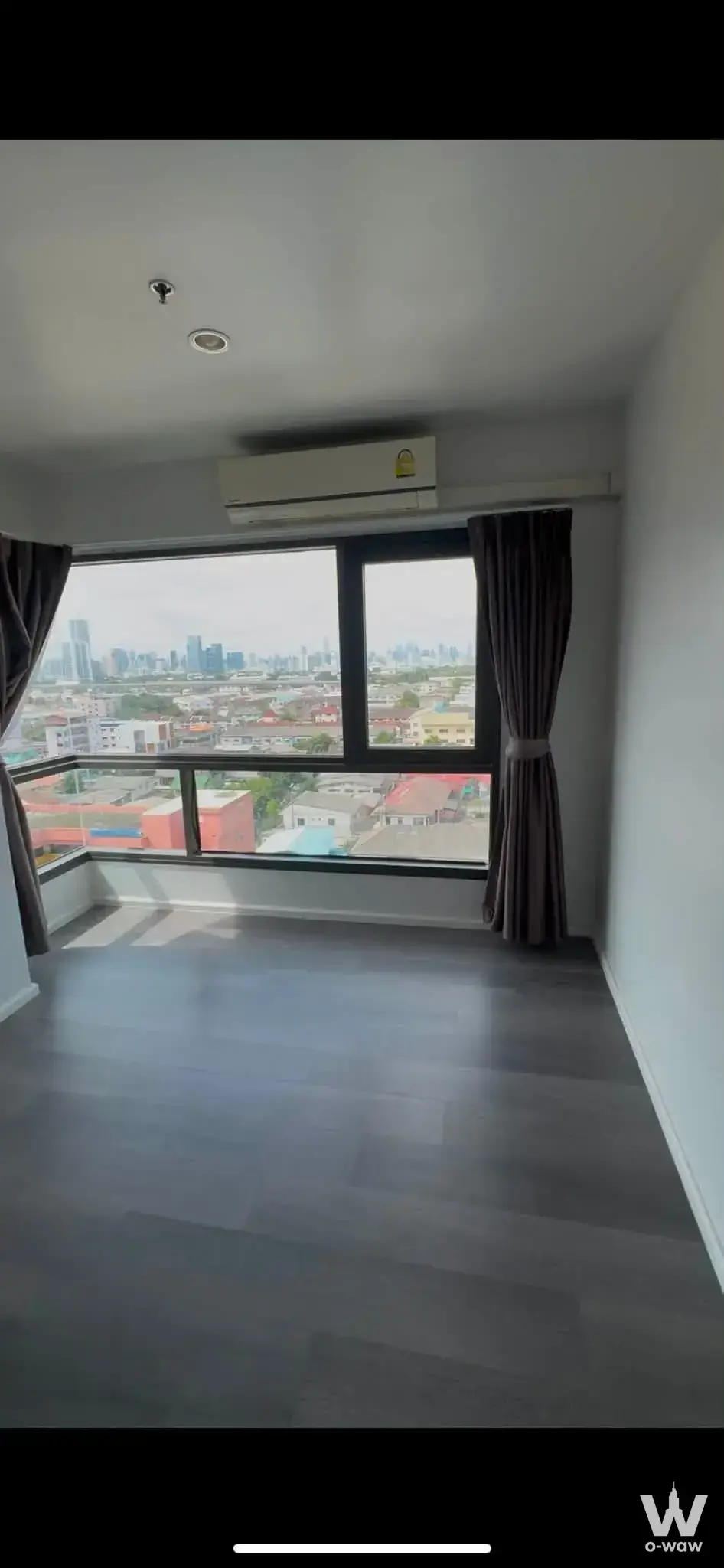 Empty modern condo room with grey flooring, floor-to-ceiling windows, and a city skyline view