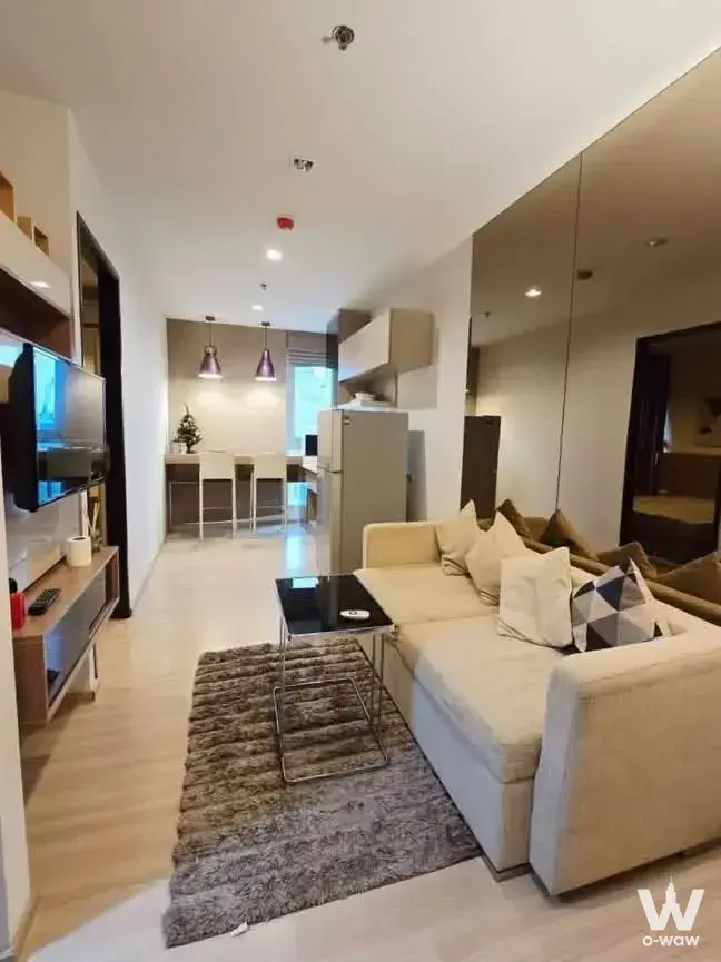 The living room has a cream sectional sofa and floor-to-ceiling mirror in a 1-bed condo for rent, RHYTHM Sathorn