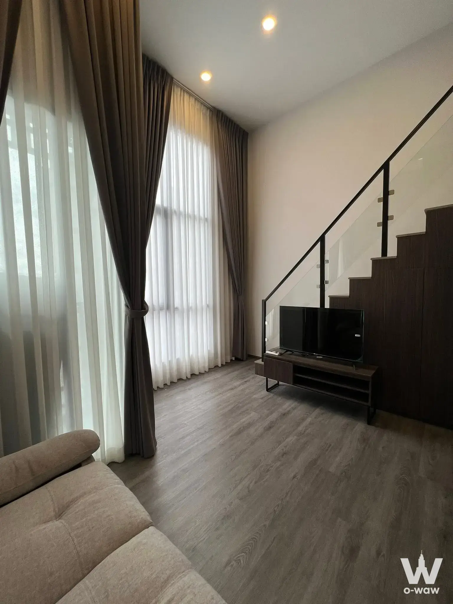 Modern living room with high ceilings, grey laminate floors, and glass staircase railing in 1-bed rental at The Origin
