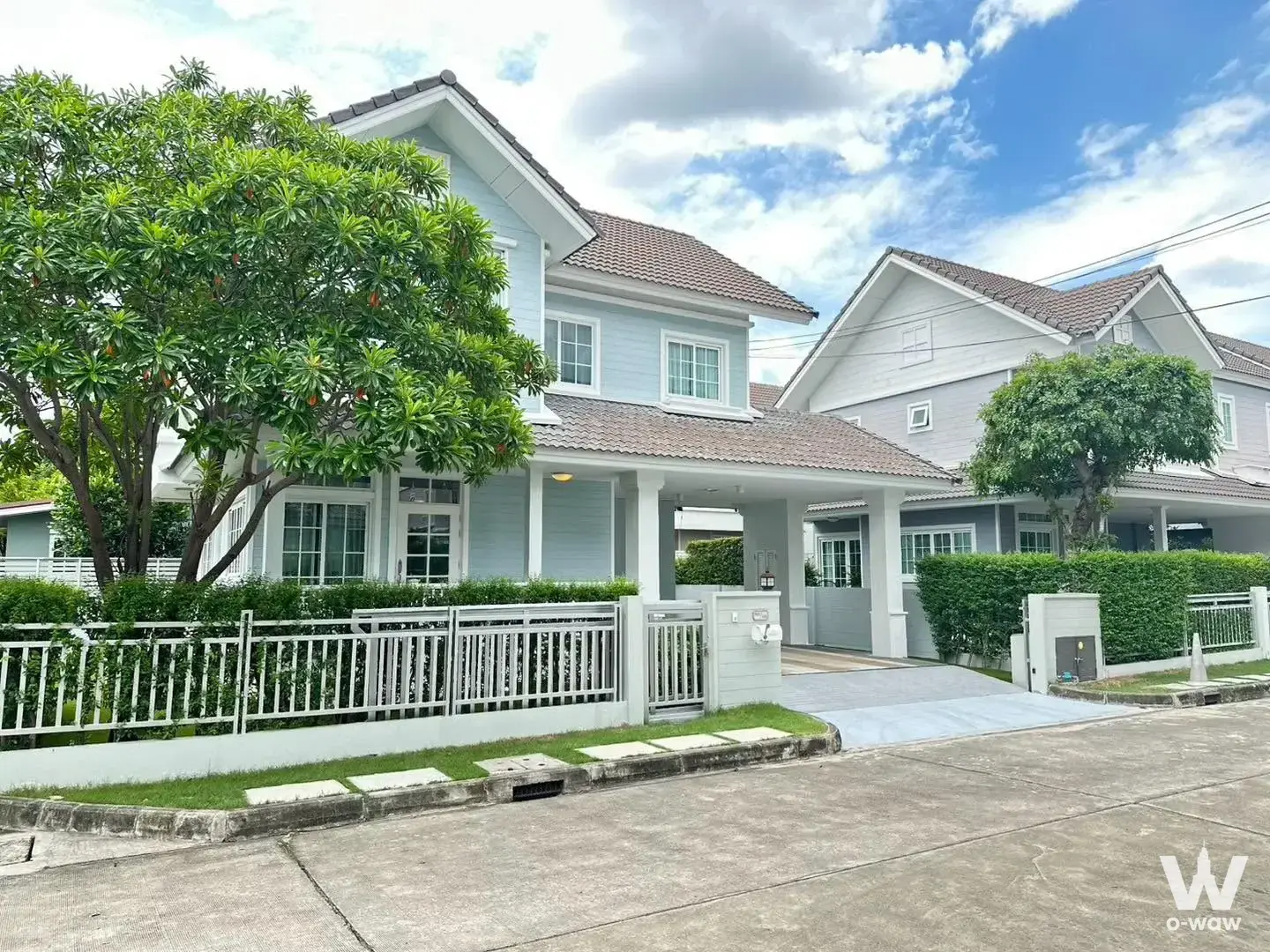 Light-blue two-story house exterior with clay tile roof and covered carport at Burasiri Panya-Indra