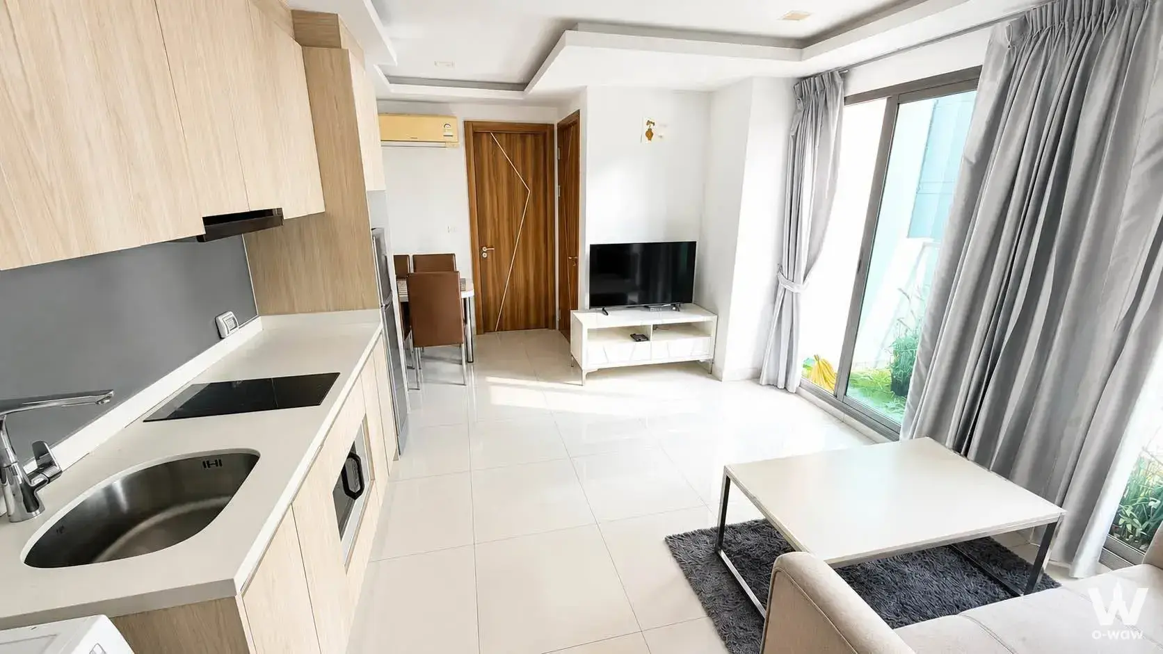 Modern open-plan living area with glossy white tiles, light wood kitchen cabinetry, and floor-to-ceiling balcony doors in