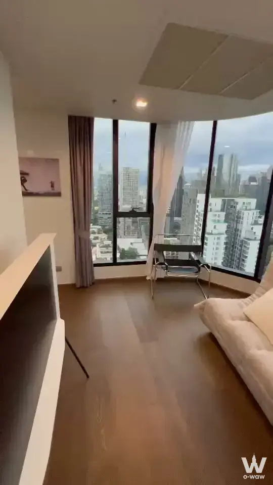 The living room in a 1-bed Ideo Q Sukhumvit 36 condo has dark wood floors, a chrome chair and curved floor-to-ceiling windows