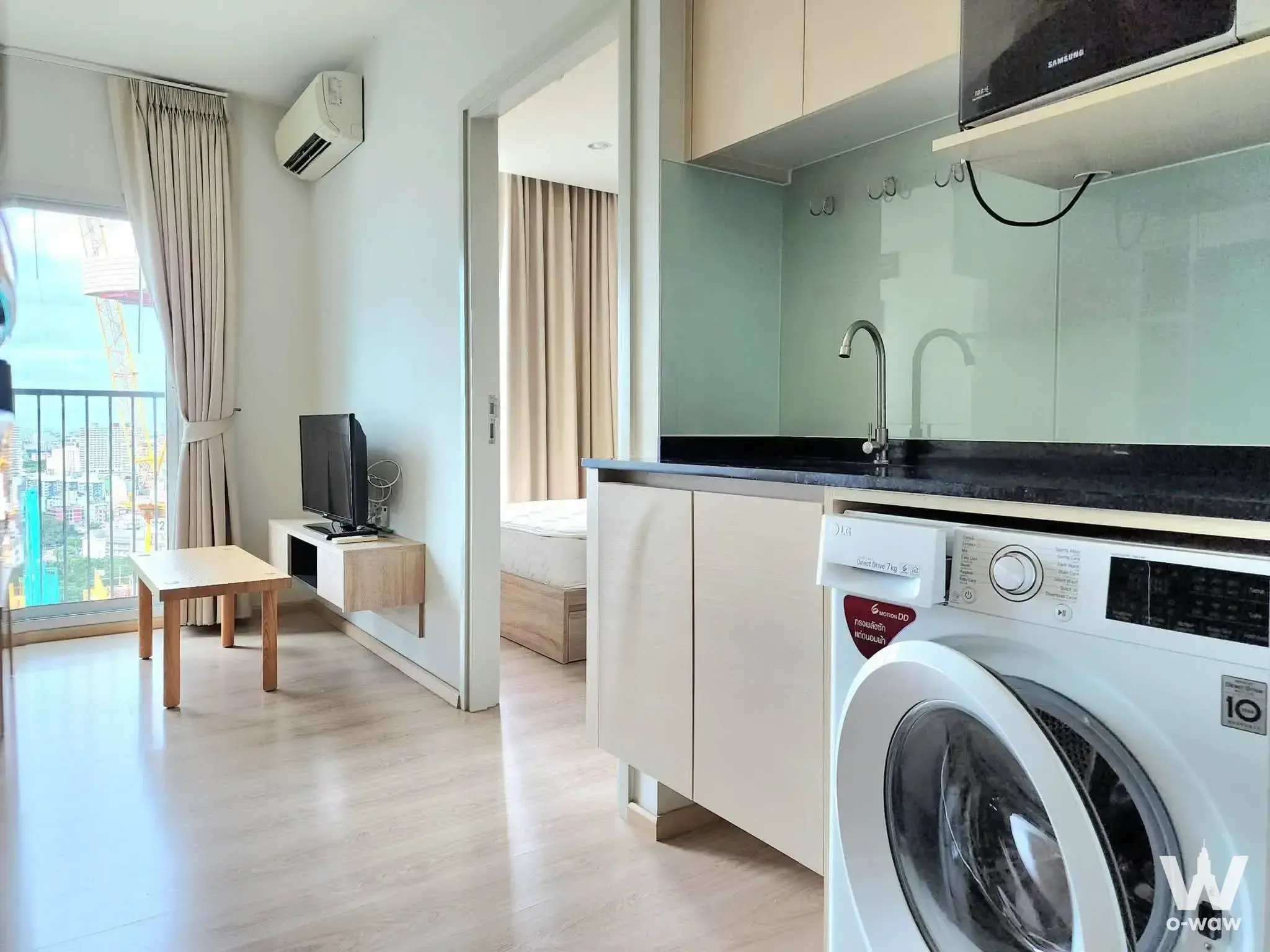 Modern studio apartment with open-plan kitchenette, light wood flooring, and floor-to-ceiling windows offering a high-rise