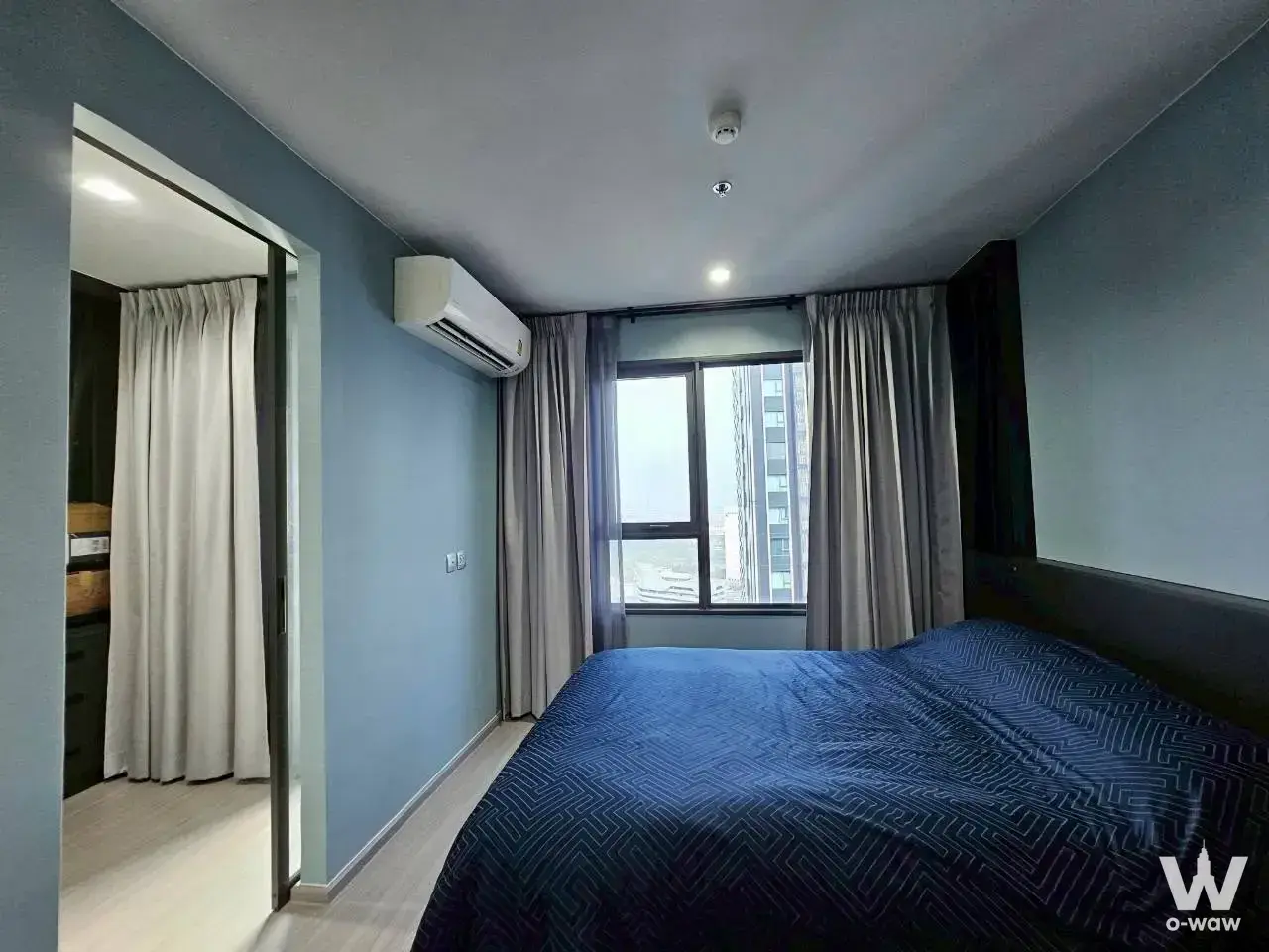 Modern bedroom in 1-bed Life Ladprao Valley condo with light blue walls, queen bed, and city view window