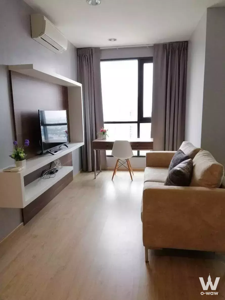 Bright 2-bed Ideo Q Ratchathewi living room with beige sofa, TV unit, and sunlit wooden desk