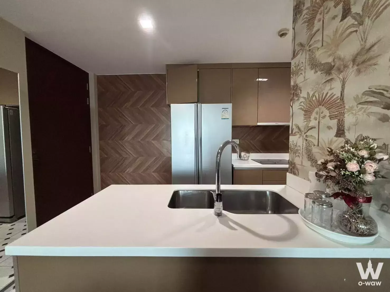 Modern kitchen with white island sink, herringbone backsplash, and tropical wallpaper in 2-bed condo for rent at Siri