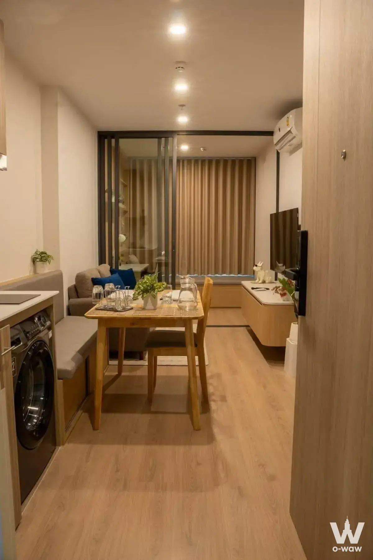 Fully furnished studio apartment featuring a wooden dining table, grey sofa, and glass-partitioned bedroom