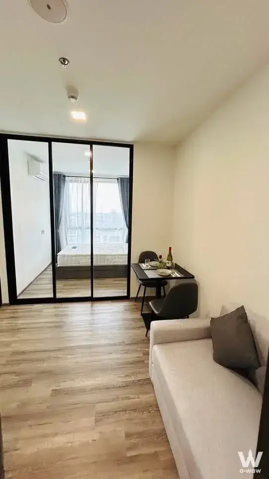 Living and bedroom combo featuring grey sofa, dining set, and glass partition revealing a queen bed