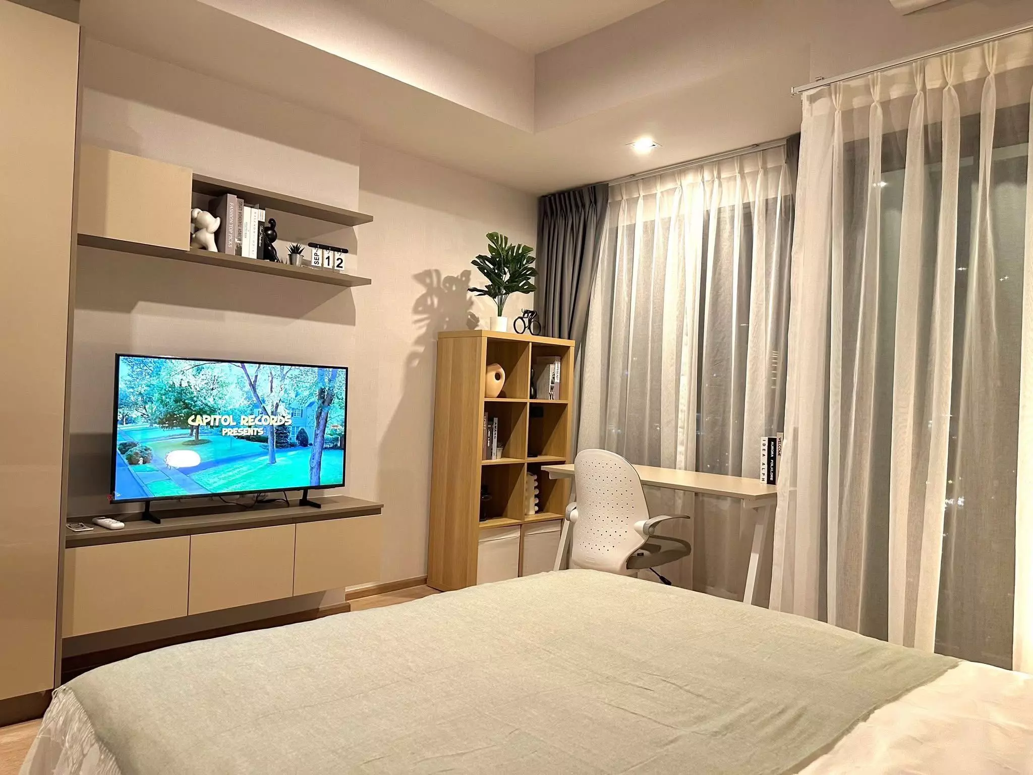 Bright bedroom in IDEO Rama 9 condo with light wood floors, built-in TV unit, and sheer curtains framing city views
