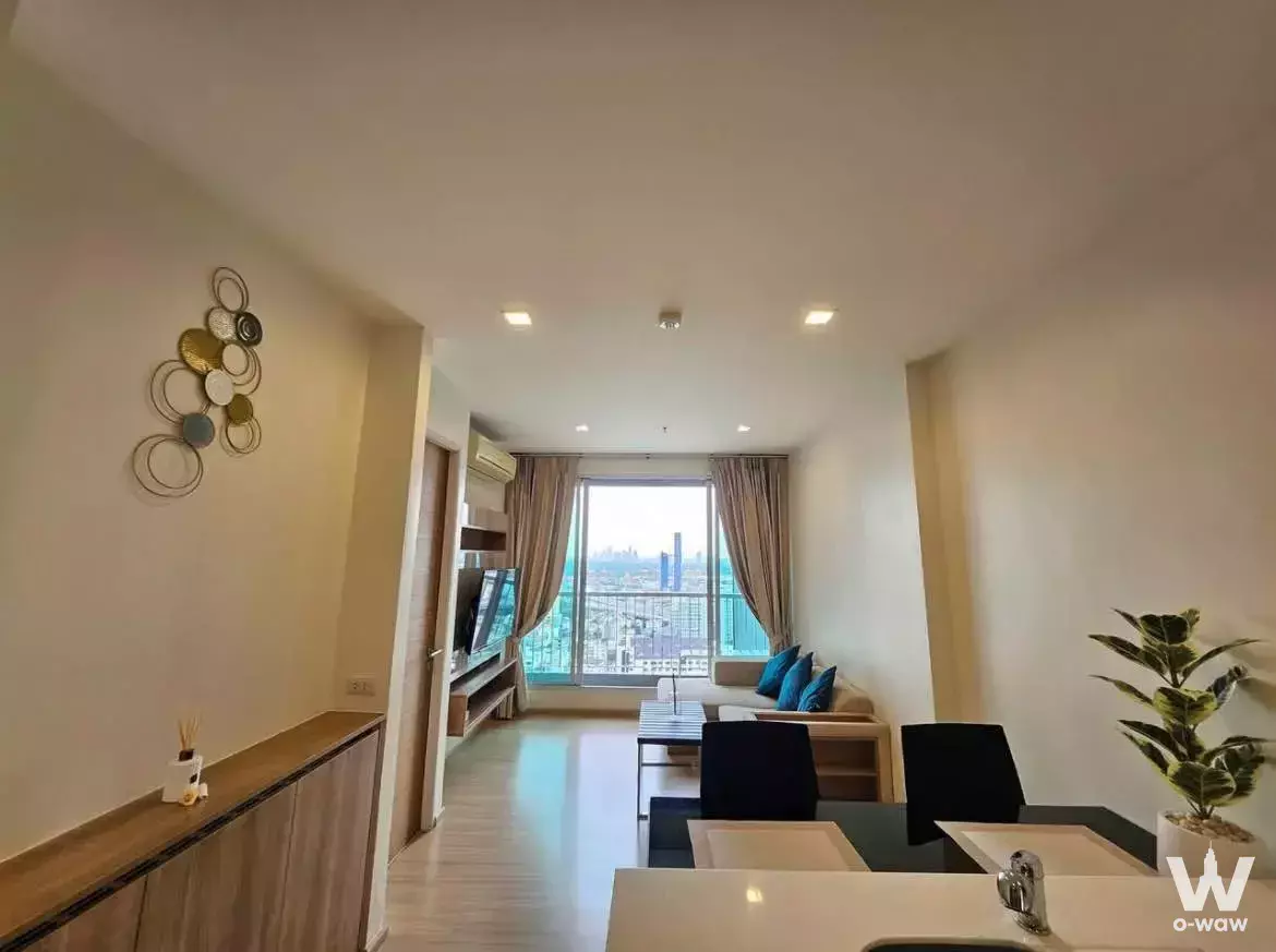 Modern 1-bed condo living area in Rhythm Sukhumvit 50 with glossy floors, white kitchen island, and city skyline views from