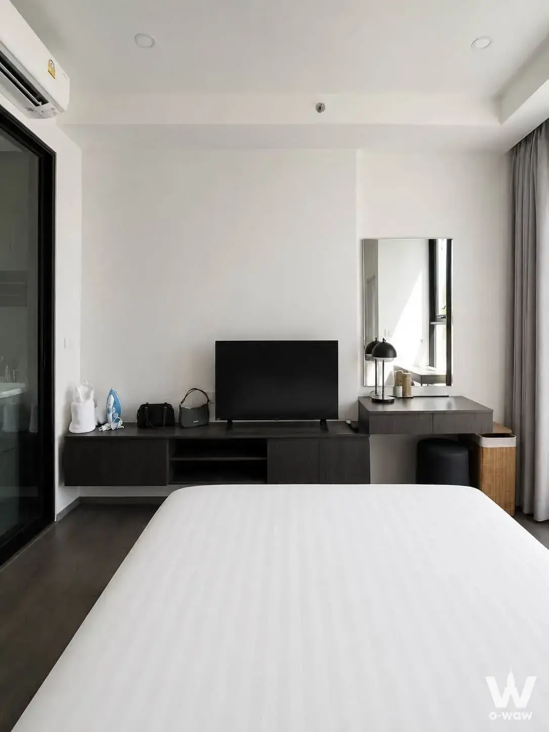 A queen bed and dark wood media console appear in a minimalist bedroom of a 1-bed condo for sale in Hampton Sriracha
