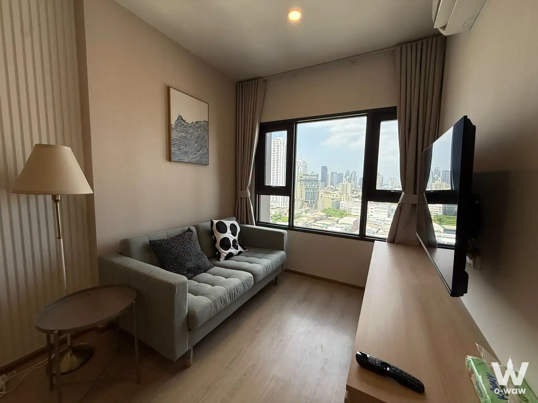 Modern studio living area with grey sofa, brass lamp, wall-mounted TV, and large windows overlooking the city skyline