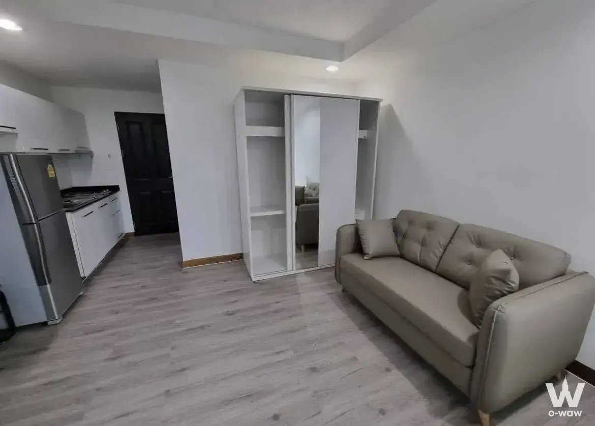 Open-plan living room in The Kris Extra 4 1-bed condo featuring taupe sofa and compact kitchenette