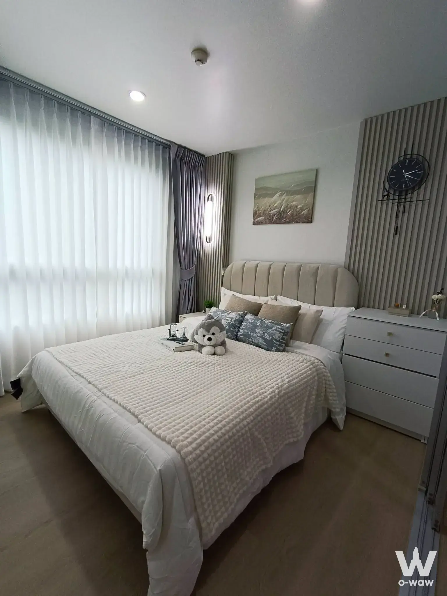 Bedroom in The Niche ID Bangkhae one-bedroom condo for sale with a beige upholstered bed, white linens and sheer curtains