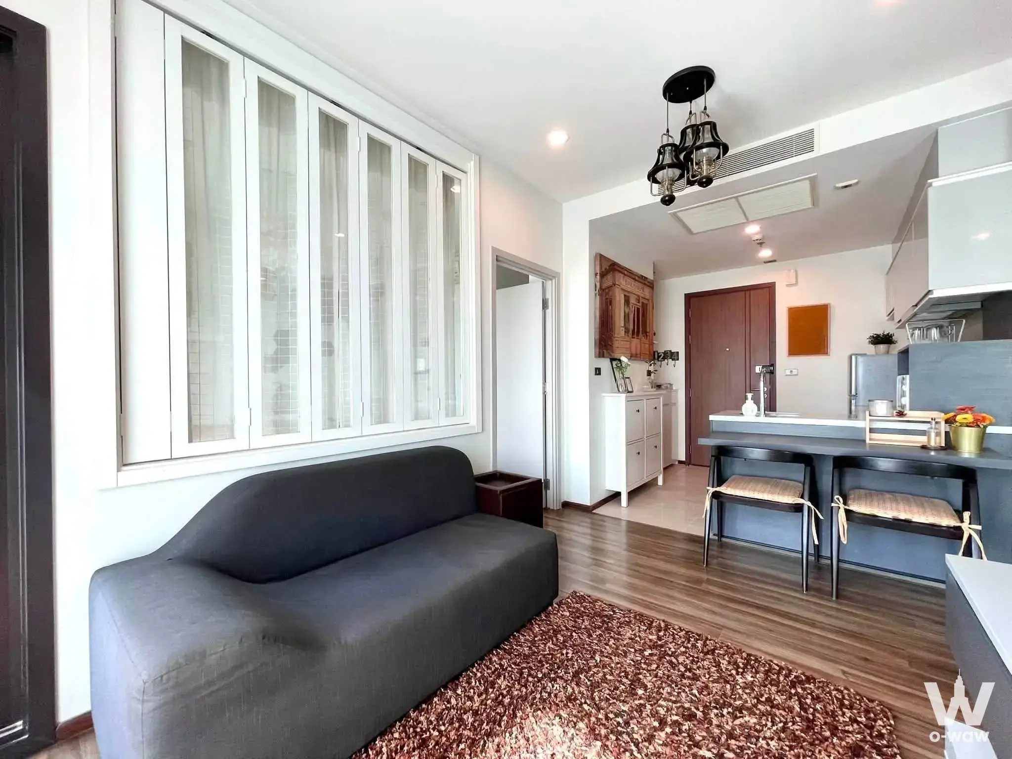 Living room and open kitchen in a WYNE by Sansiri 1-bed condo for sale with a grey sofa, breakfast bar and large windows