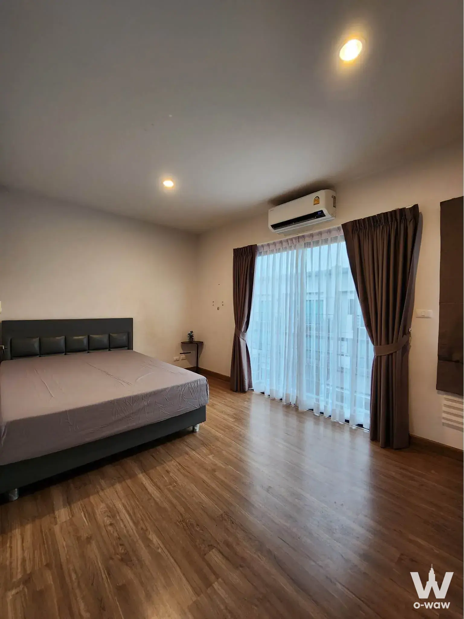 Minimalist bedroom in Baan Klang Mueang townhouse with grey upholstered bed, wood-laminate floors, and floor-to-ceiling