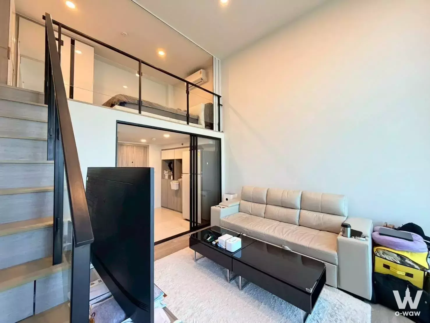 Loft living area in Origin Nonthaburi with beige sofa, mezzanine bed, and open-plan kitchen under high ceilings