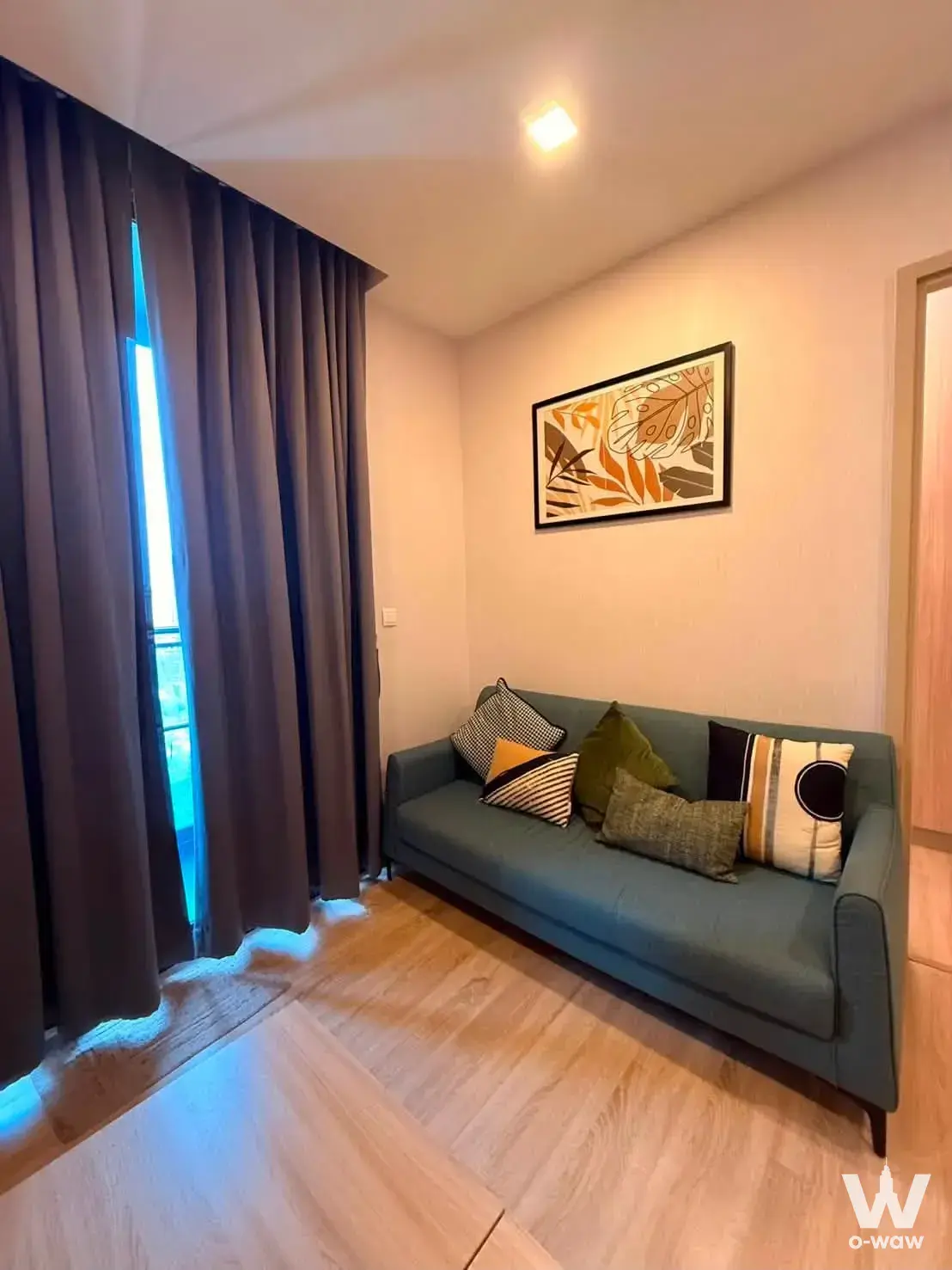 Compact living space with teal sofa, patterned pillows, botanical art, and balcony access through curtains
