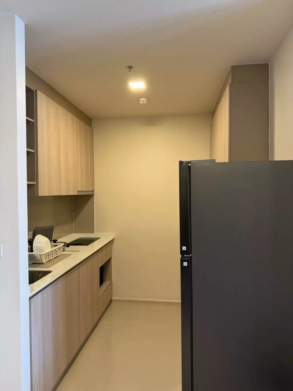 Sleek galley kitchen with light wood cabinets, black appliances, and cream tiles in this FLO by Sansiri rental unit