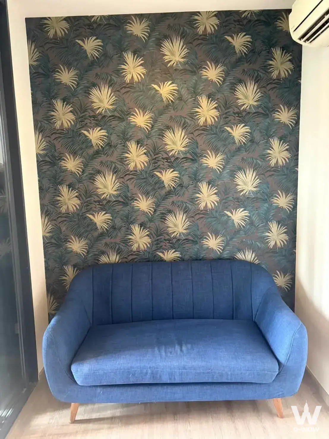 Living room corner in THE LINE Wongsawang featuring a blue tufted sofa against tropical palm wallpaper