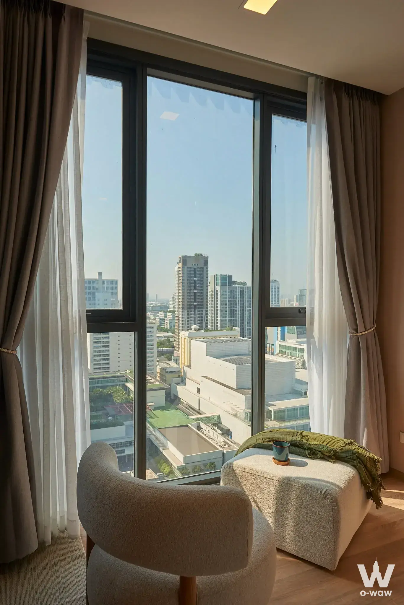 Living corner in an XT Phayathai condo featuring floor-to-ceiling windows, city views, and a cream boucle armchair