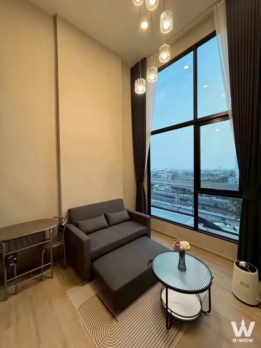 Living room in Origin Plug & Play condo featuring grey sofa, crystal pendants, and city skyline views at dusk