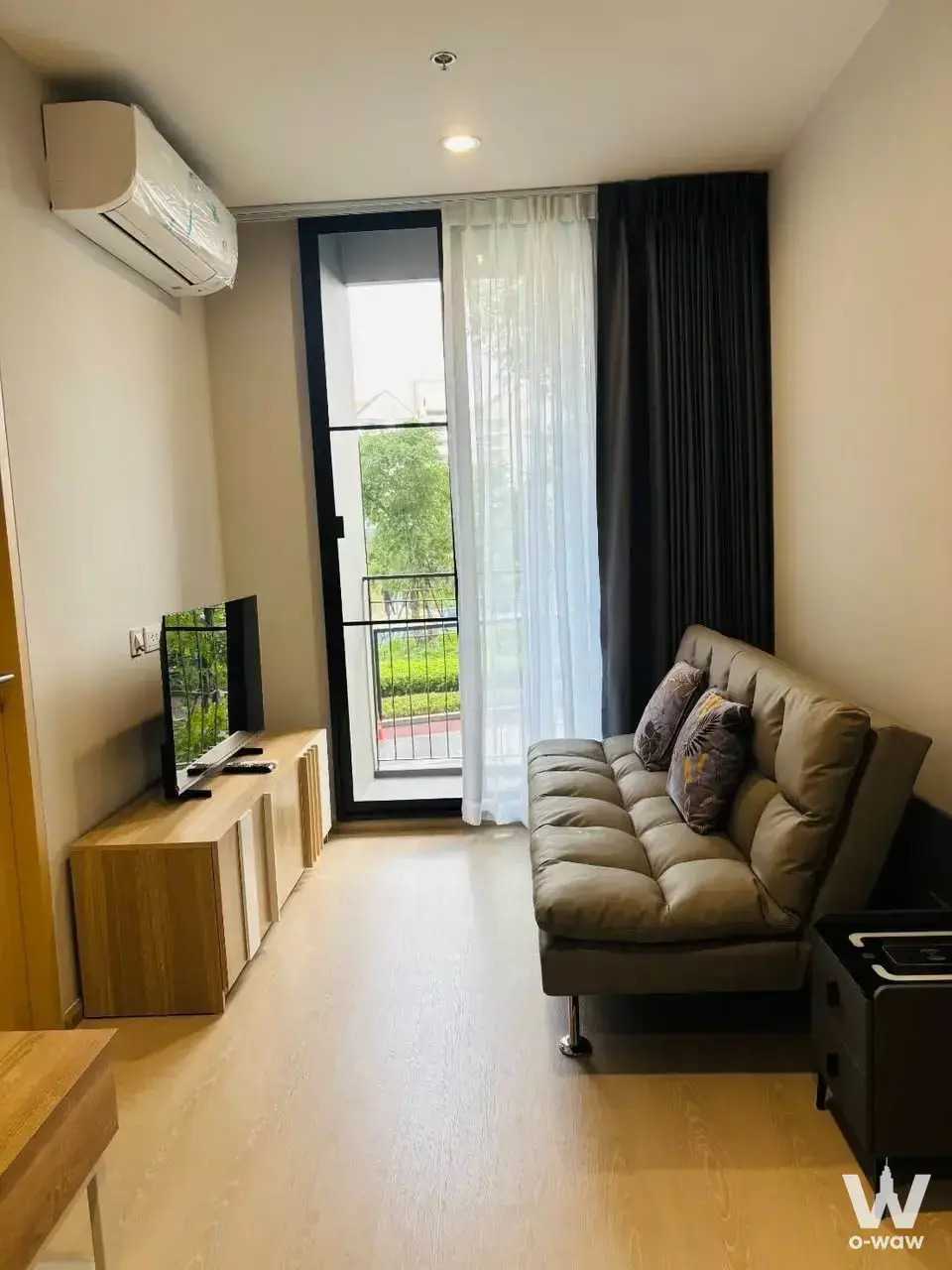 Skyrise Avenue Sukhumvit 64 living room with taupe sofa, light-wood console, and balcony access