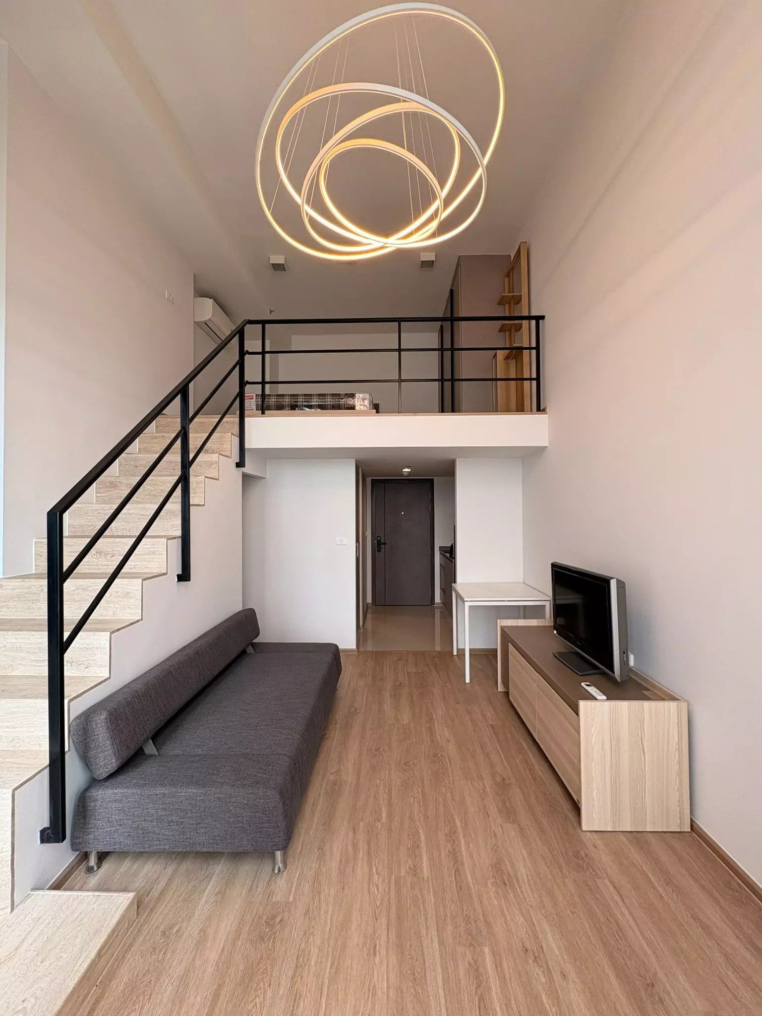 High-ceiling duplex living area in IDEO Rama 9 condo with floating staircase to mezzanine loft, grey futon sofa, and