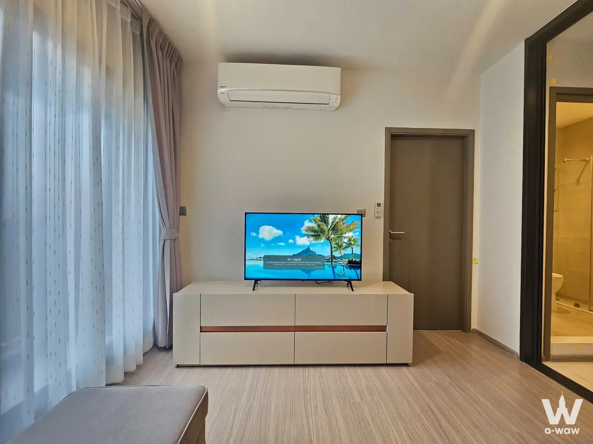 Modern living room with light wood floors, white media console, TV, and glass door to bathroom
