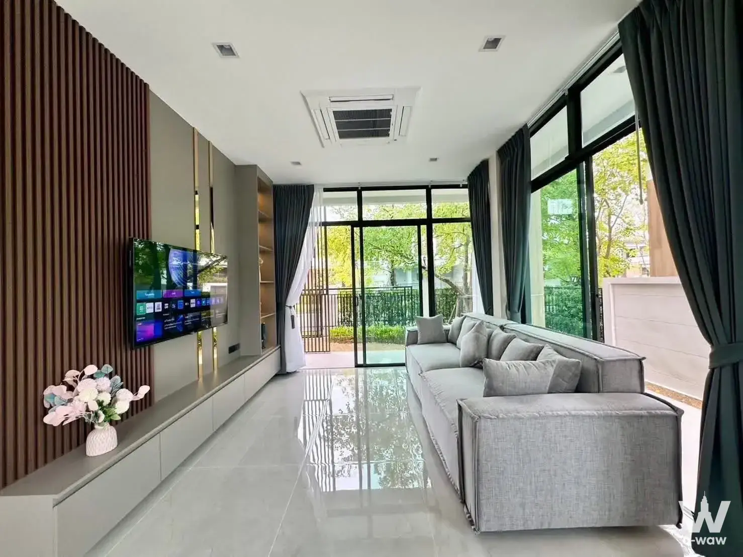 Living room in 4-bed Bangkok house with L-shaped grey sofa, wood slat accent wall, and floor-to-ceiling windows