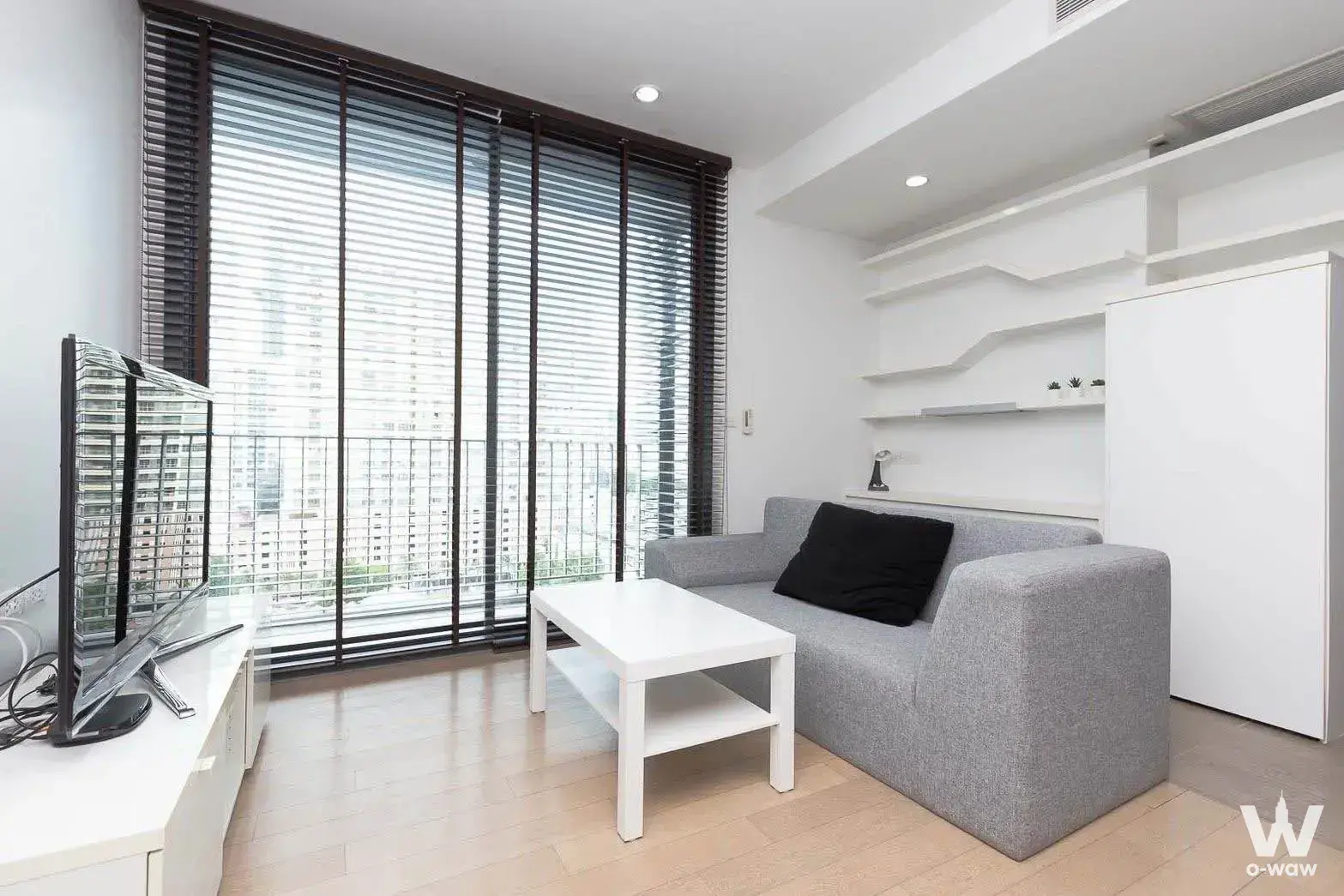 Minimalist living room in a PYNE by Sansiri 1-bed condo with grey sofa, wooden blinds, and built-in white shelving